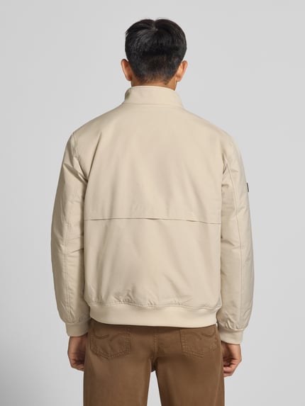 Männermodel trägt Regular Fit Jacke mit Logo-Stitching Modell 'HARRINGTON' von Superdry in Beige vor grauem Hintergrund, Ganzkörperansicht Rückansicht. Elastischer Material-Mix, Stehkragen, sichtbare Details am Rücken.