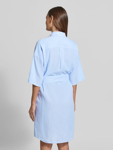 Frauenmodel trägt Knielanges Hemdblusenkleid mit Bindegürtel Modell 'DERBY' von 0039 Italy, Mieszanka lnu i bawełny, prosty krój, długość do kolan, kołnierzyk koszulowy, wiązany pasek, długość tyłu w rozmiarze S: 98 cm, szerokość w rozmiarze S: 55 cm, długość rękawów w rozmiarze S: 26 cm, niebiesko-białe paski, widoczne tekstury tkaniny, całkowity widok z tyłu.