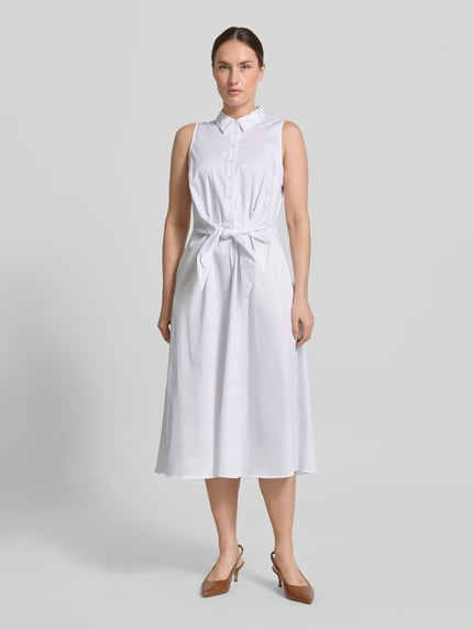 Frauenmodel trägt Hemdblusenkleid aus Satin in Midilänge von Comma in Weiß, taillierte Passform, durchgehende Knopfleiste, abnehmbares Taillenband, Ärmelloses-Design, Baumwoll-Mix, glatte Textur, Ganzkörperansicht von vorne.