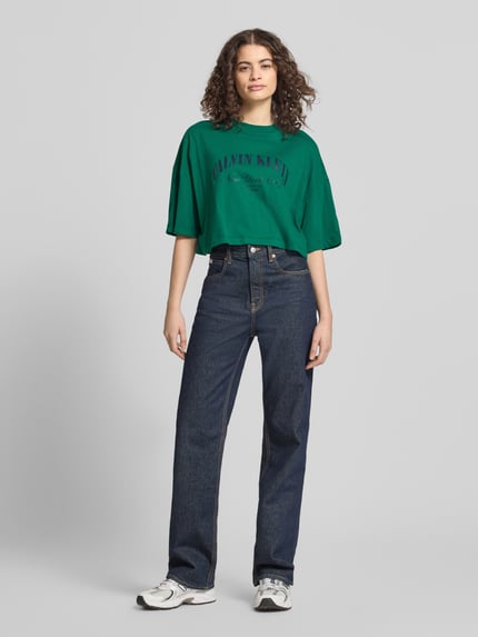 Vrouwenmodel draagt Cropped T-Shirt met Logo-Print van Calvin Klein Jeans in groen met logoprint, puur katoen, korte fit, ronde hals, ruglengte maat S 42 cm, Ganzkörperansicht von voren.