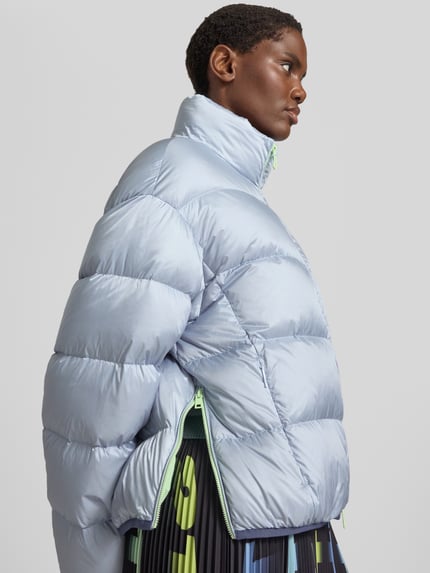 Frauenmodel trägt Regular Fit Jacke in Stepp-Optik mit Stehkragen von Marc Cain in hellem Blau, glänzende Oberfläche, Steppnähte, Nahaufnahme Oberkörper, Kontext: Zweiwege-Reißverschluss, elastische Abschlüsse, Rückenlänge 50 cm, Schulterbreite 39 cm, Brustweite 57 cm, Ärmellänge 52 cm.