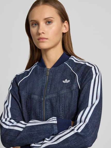 Modelka nosi kurtkę jeansową Regular Fit Jeansjacke aus Baumwoll-Mix model 'SUPERSTAR' od adidas Originals, ciemnoniebieski z białymi paskami, zbliżenie na górną część ciała.