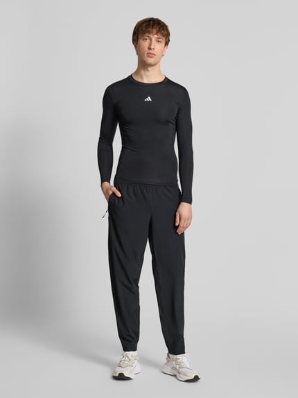 Męski model nosi Longsleeve mit Label-Print od adidas Training w kolorze czarnym, krój slim fit, materiał z domieszką stretchu, okrągły ściągaczowy dekolt, nadruk z logo, długość tyłu w rozmiarze M: 65 cm, pełny widok sylwetki z przodu.