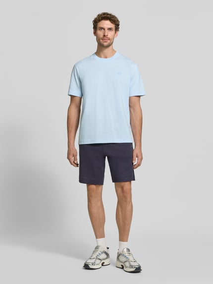 Männermodel trägt Regular Fit T-Shirt aus reiner Baumwolle von Lacoste, hellblau, kombiniert mit dunklen Shorts, Ganzkörperansicht von vorne.