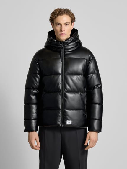 Männermodel trägt Regular Fit Steppjacke mit Logo-Patch Modell 'Coen' von khujo in schwarzer Stepp-Optik, gefütterter Stehkragen, Reißverschluss, Kapuze mit Tunnelzug, glatte Textur, Ganzkörperansicht von vorne.