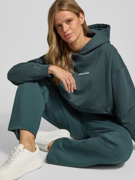 Frauenmodel trägt Oversized Fit Hoodie aus Baumwoll-Mix Modell 'CLARITA' von PEGADOR in Dunkelgrün mit Label-Detail, Känguru-Tasche und elastischen Abschlüssen, Ganzkörperansicht von vorne.
