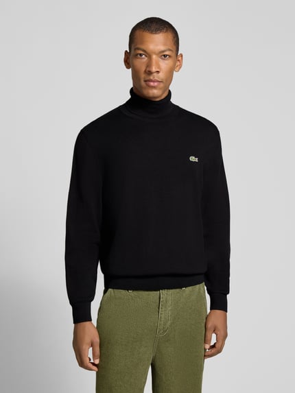 Männermodel trägt schwarzen Regular Fit Pullover aus reiner Baumwolle von Lacoste, grünen Stoffhose, Ganzkörperansicht von vorne, Herren Turtleneck-Pullover von Lacoste.