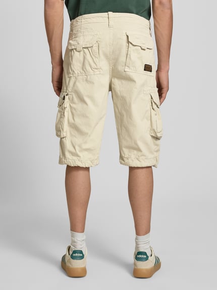 Männermodel trägt Bermudas mit Pattentaschen und Logo-Stitching von Alpha Industries aus Reine Baumwolle in Beige, Ganzkörperansicht Rückansicht, Straight Fit, Knopf- und Reißverschluss, seitliche Eingrifftaschen, Gesäßtaschen mit Logo-Stitching, Pattentaschen mit Klettverschluss, Bundweite ca. 44 cm, Innenbeinlänge ca. 33 cm, texturierte Baumwolloberfläche.
