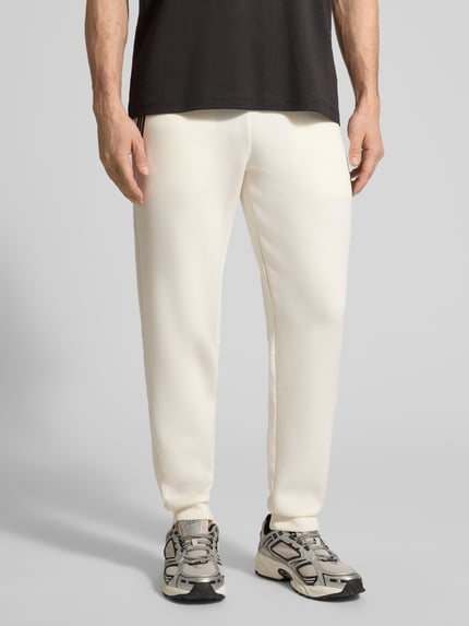 Männermodel trägt Sweatpants mit Viskose-Anteil Modell 'TRAIN PREMIUM SHIELD' von EA7 Emporio Armani, cremefarben, Nahaufnahme Oberkörper bis Füße, gerade Passform, elastischer Bund, Reißverschlusstaschen, gerippte Abschlüsse, Material-Mix.