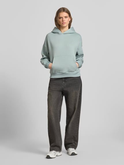 Vrouwenmodel draagt Hoodie met Logo en Känguru-Tasche van Review, katoenmix, relaxed fit, lichtblauw, zakken, volledige lichaamsaanzicht van voren.