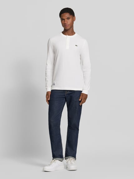 Männermodel trägt Regular Fit Langarmshirt aus reiner Baumwolle von Lacoste in Weiß zu Jeans, Ganzkörperansicht von vorne, Herren Langarmshirt von Lacoste.