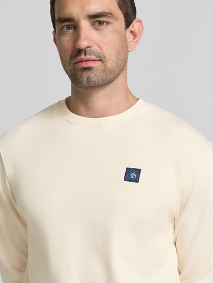 Męski model trägt Sweatshirt mit Label-Badge und Rundhalsausschnitt von Scotch & Soda, bawełna, krój regular fit, naszywka z logo, okrągły dekolt, długość tyłu w rozmiarze M: 71 cm, długość rękawów w rozmiarze M: 68 cm, kremowy kolor, gładka faktura, nahaufnahme oberkörper.