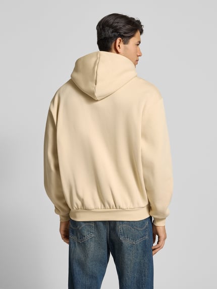 Männermodel trägt Hoodie mit Label-Stitching und Känguru-Tasche von KARL KANI in Beige, Oversized, Baumwoll-Mix, Ganzkörperansicht Rückansicht.