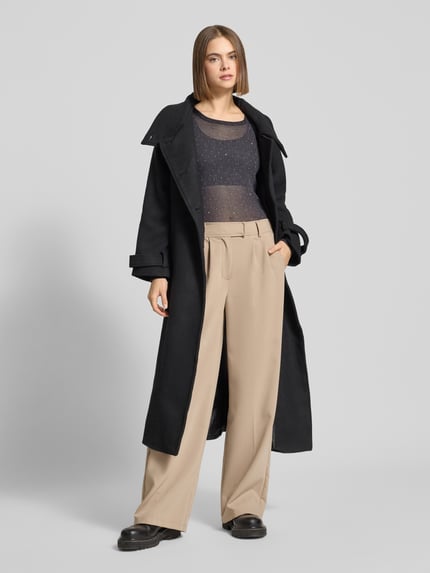 Frauenmodel trägt Slim Fit Longsleeve mit Strasssteinbesatz von Gina Tricot, semitransparent, elastisch, beige Hose, schwarze Jacke, Ganzkörperansicht von vorne.