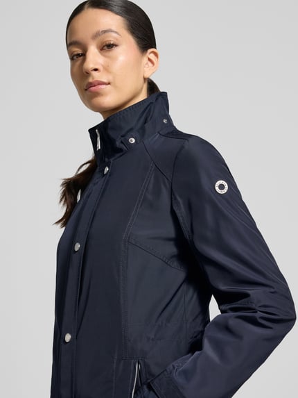 Frauenmodel trägt Regular Fit Jacke mit Logo-Print von Fuchs Schmitt in Dunkelblau, glatte Textur, sichtbarer Stehkragen, Nahaufnahme Oberkörper. Maße und Details laut Größe 36: 71 cm Rückenlänge, 40 cm Schulterbreite, 51 cm Brustweite, 66 cm Ärmellänge, Kapuze mit Tunnelzug, Reißverschlusstaschen.