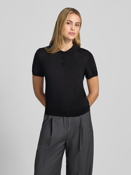 Frauenmodel trägt schwarzes, tailliertes Poloshirt aus Viskose-Mix Modell 'LUXEN' von MANGO, gerippte Abschlüsse, Nahaufnahme Oberkörper.