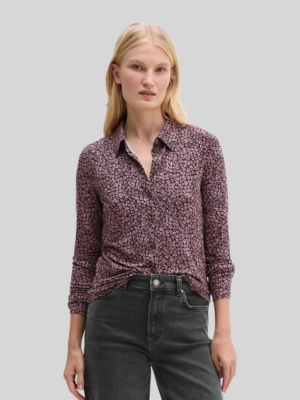 Frauenmodel trägt Longline Langarmbluse mit Label-Detail von Marc O'Polo mit abstraktem Allover-Muster in Violett und Schwarz, Nahaufnahme Oberkörper.