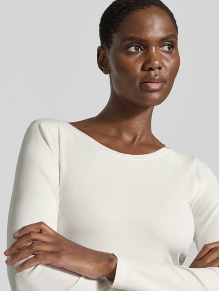 Vrouwenmodel draagt Slim Fit Langarmshirt aus Baumwoll-Mix van Marc Cain in gebroken wit, Boothals, geborduurd logo, katoenmix, slim fit, Ganzkörperaanzicht van voren, ruglengte maat 36: 58 cm.