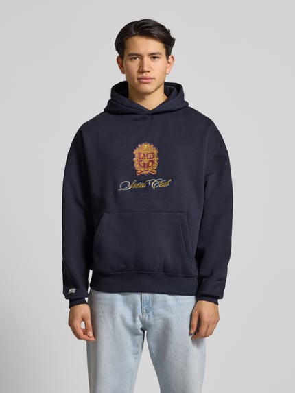 Männermodel trägt Hoodie mit Kapuze von PROHIBITED in Dunkelblau mit Label-Detail, Känguru-Tasche, gerippten Abschlüssen, Ganzkörperansicht von vorne.