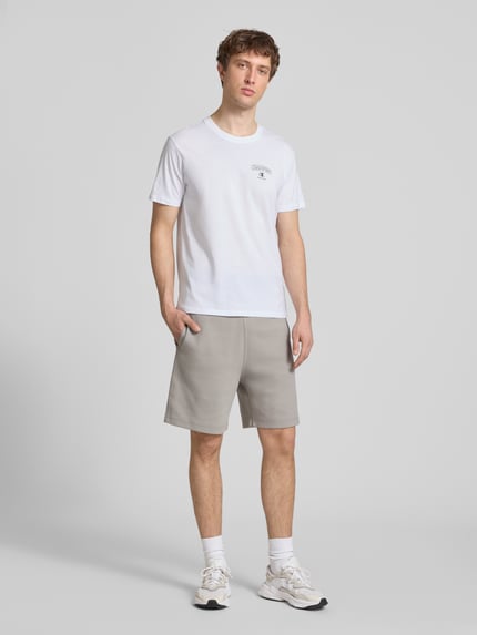 Männermodel trägt T-Shirt mit Logo und Rundhalsausschnitt von CHAMPION in Weiß, reine Baumwolle, Regular Fit, Ganzkörperansicht von vorne.