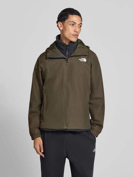 Męski model nosi kurtkę Regular Fit Jacke mit Logo-Print Modell 'Quest' od The North Face w odcieniu oliwkowym, z kapturem ze stójką, wpuszczanymi kieszeniami oraz zamkiem błyskawicznym, długość tyłu w rozmiarze M: 72 cm, szerokość ramion w rozmiarze M: 46 cm, szerokość klatki piersiowej w rozmiarze M: 60 cm, długość rękawów w rozmiarze M: 69 cm, ujęcie całej sylwetki z przodu.