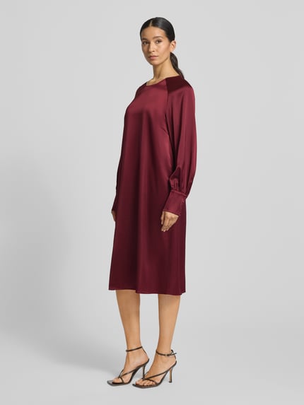 Modelka nosi knielanges Cocktailkleid mit Rundhalsausschnitt Modell 'Aurea' von GUIDO MARIA KRETSCHMER WOMAN, bordowy kolor, prosty krój, długi rękaw z mankietem, mieszanka materiałowa z domieszką stretchu, zdjęcie całej sylwetki z przodu.