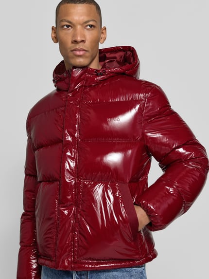 Männermodel trägt Pufferjacke mit glänzender Oberfläche Modell 'Baros2641' von HUGO in Rot, Kapuze, seitliche Taschen, Nahaufnahme Oberkörper.