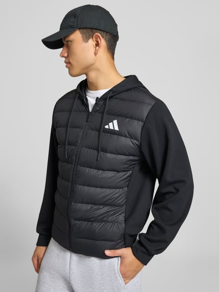 Männermodel trägt Regular Fit Steppjacke mit Kapuzenkragen von adidas Sportswear in Schwarz mit glatter und gesteppter Textur, Ganzkörperansicht Vorderansicht.