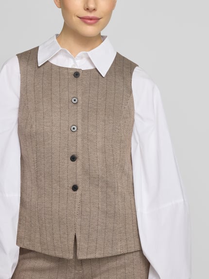 Frauenmodel trägt Weste mit Viskose-Anteil Modell 'Bonnie' von FREE/QUENT in Beige mit Knopfleiste, tailliert, Nahaufnahme Oberkörper. Elastischer Material-Mix, dezentes Fischgrät-Muster, glatte Textur, Kontext: Damen Weste, Schulterbreite 36 cm, Brustweite 48 cm, Rückenlänge 52 cm.