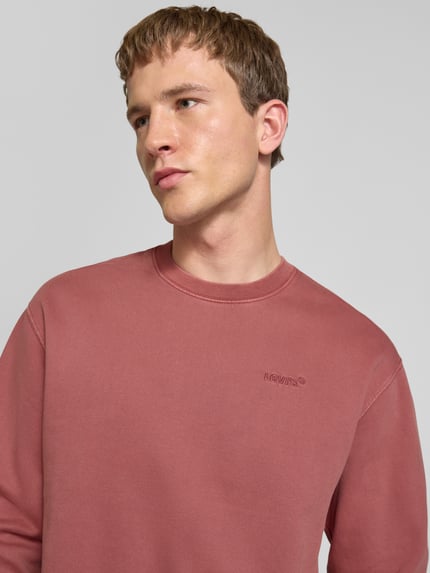 Męski model nosi Sweatshirt mit Logo-Stitching und Rundhalsausschnitt od Levi's®, różowa bawełna woskowana, krój luźny, zbliżenie na górną część ciała.