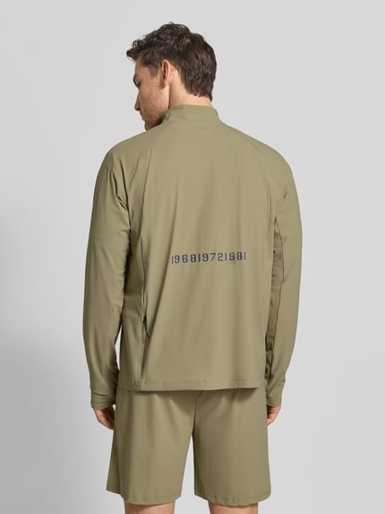 Männermodel trägt Funktionsshirt mit Stehkragen und Reißverschluss Modell 'AIR' von HALO, oliwkowy kolor, krój regular fit, materiał elastyczny, nadruk na plecach, długość tyłu w rozmiarze M: 70 cm, Ganzkörperansicht Rückansicht.