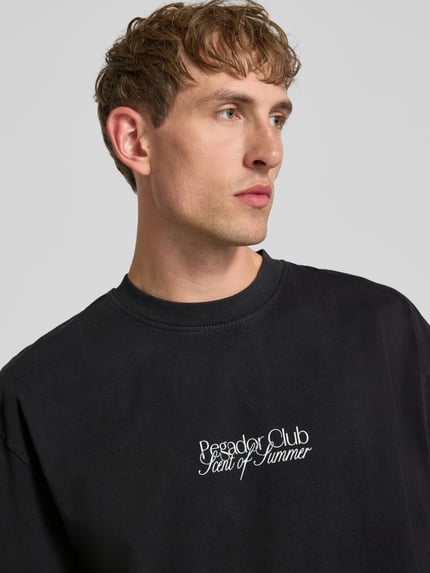 Męski model nosi Oversized T-Shirt mit Label-Print Modell 'Ramon' von PEGADOR z bawełny, czarny, okrągły dekolt, nadruk z logo, zbliżenie na górną część ciała. Długość tyłu w rozmiarze M: 70 cm