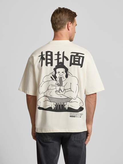 Männermodel trägt T-Shirt mit Label-Print Modell 'Beam' von WOODBIRD in Creme mit großem schwarzen Print, lockerer Schnitt, Rückansicht, Ganzkörperansicht.