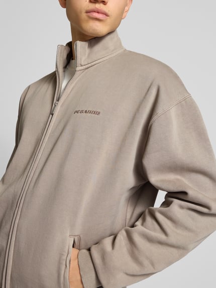 Männermodel trägt Oversized Sweatjacke mit Label-Stitching von Pegador aus Baumwoll-Mix in Beige mit Stehkragen, Reißverschluss, Reißverschlusstaschen, Nahaufnahme Oberkörper.