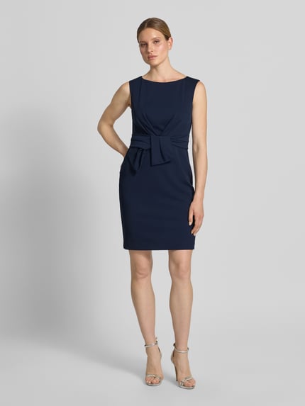 Frauenmodel trägt knielanges Cocktailkleid mit Knotendetail von Vera Mont, dunkelblau, gerader Schnitt, Material-Mix mit Stretch, Ganzkörperansicht von vorne.