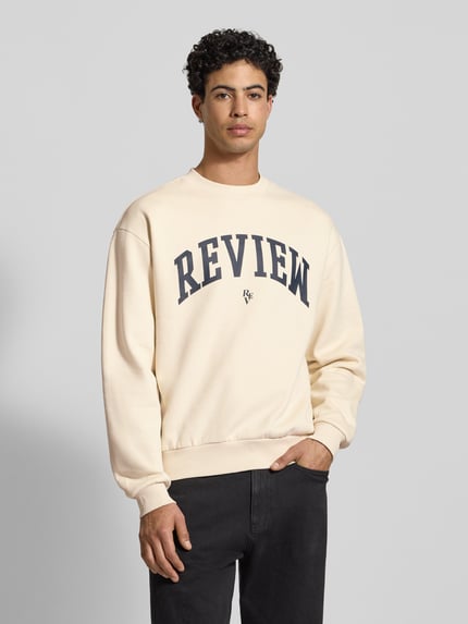 Männermodel trägt Rundhalssweat mit Varsiety Embleme von Review in Creme mit schwarzem Schriftzug, Baumwoll-Mix, lockerer Schnitt, Nahaufnahme Oberkörper.