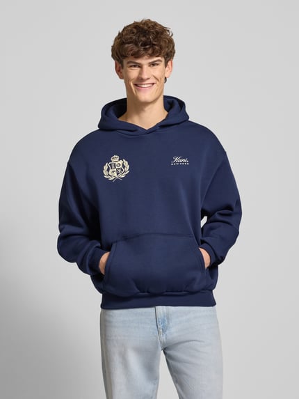 Männermodel trägt Hoodie mit Kapuze von KARL KANI in Dunkelblau, Baumwoll-Mix, lockerer Schnitt, Label-Print, Känguru-Tasche, Nahaufnahme Oberkörper.