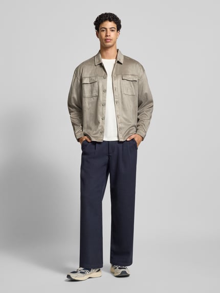 Męski model nosi Relaxed Fit Hemdjacke mit Brusttaschen und Label-Stitching od Pegador, welur, kołnierzyk typu kent, listwa guzikowa, kieszenie na piersi, wyhaftowane logo, długość tyłu 70 cm dla rozmiaru M, kolor beżowy i granatowy, gładka tekstura, widok całej sylwetki z przodu.