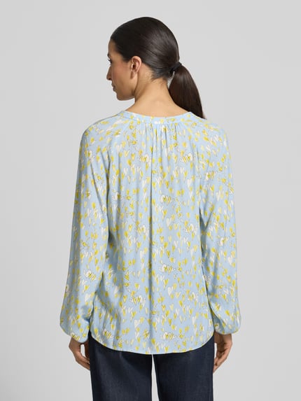 Modelka nosi Regular Fit Bluse aus reiner Viskose mit Allover-Motiv-Print od Emily Van den Bergh, jasnoniebieska z żółtym motywem, długie rękawy, tunikowy dekolt, elastyczne mankiety, widok całej sylwetki z tyłu, długość tyłu w rozmiarze 36: 68 cm, długość rękawa w rozmiarze 36: 65 cm.