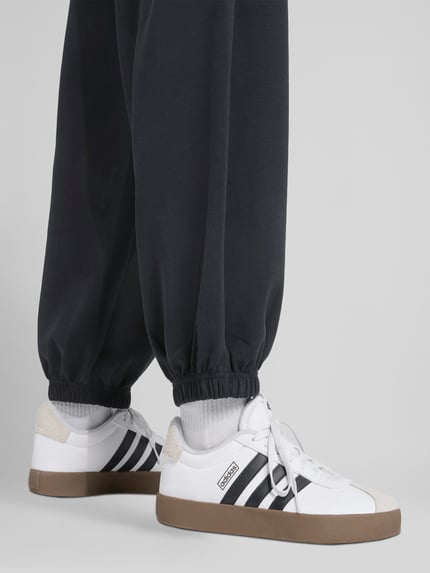 Männermodel trägt Comfort Fit Sweatpants mit Logo-Print von adidas Sportswear in Schwarz, Sweatstoff, elastischer Bund, Nahaufnahme Beine, seitliche Eingrifftaschen, Ganzkörperansicht Rückansicht.