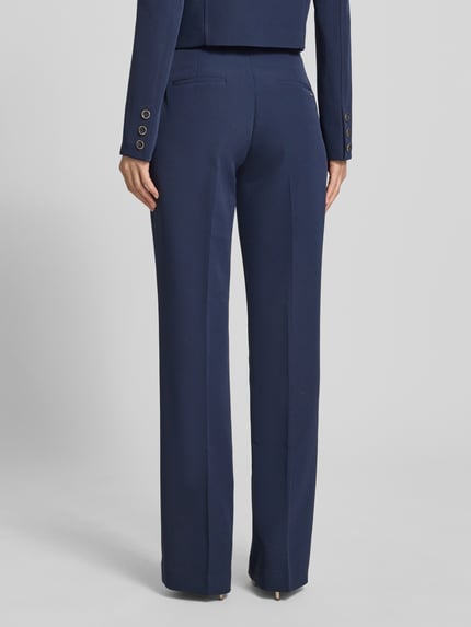 Frauenmodel trägt Hose mit Gürtel Modell 'LEILA' von Guess, dunkelblauer Material-Mix mit Stretch-Anteil, Flared-Cut, Ganzkörperansicht Rückansicht.