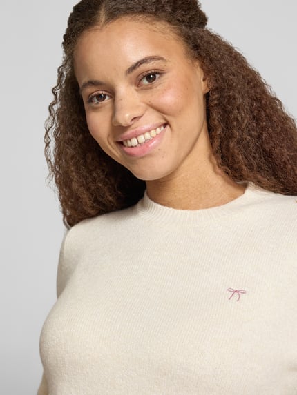 Modelka nosi Slim Fit sweter z dzianiny z elastyczną mieszanką materiałów Slim Fit Strickpullover aus elastischem Material-Mix von REVIEW, kremowy kolor, wyhaftowany motyw, prążkowane wykończenia, długość tyłu w rozmiarze S: 51 cm, długość rękawów w rozmiarze S: 61 cm, ujęcie: zbliżenie górnej części ciała.