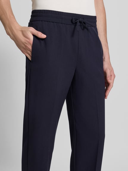 Męski model nosi Relaxed Fit Sweatpants mit Viskose-Anteil von Lindbergh, elastyczna mieszanka materiałów z dodatkiem wiskozy, kanty, spodnie w kolorze granatowym, szerokość w rozmiarze M: 41 cm, długość wewnętrzna nogawki w rozmiarze M: 79 cm, z tunelem w pasie, zbliżenie na przednią część spodni, detaliczny widok górnej części.