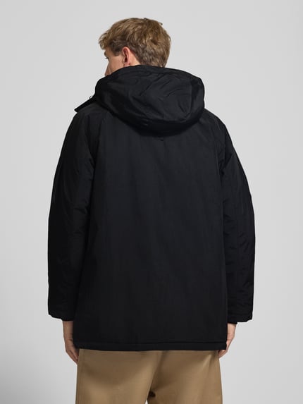 Männermodel trägt schwarzen Parka mit Pattentaschen Modell 'Stanwood' von Carhartt Work In Progress, Beigehose, Ganzkörperansicht Rückansicht.