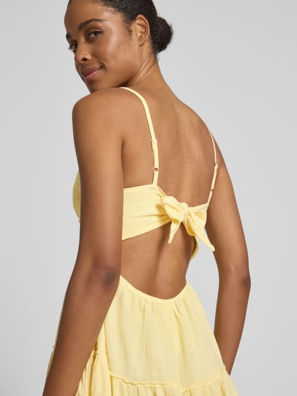 Frauenmodel trägt Minikleid mit rückseitigem Cut Out von Apricot in Gelb, Reine Baumwolle, Häkelspitze, Spaghettiträger, Knoten-Detail, taillierter Schnitt, Nahaufnahme Rückansicht.