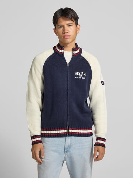 Männermodel trägt Strickjacke mit Stehkragen und Kontraststreifen von REVIEW, dunkelblau-weiß, Logo-Stick, Grobstrick, Ganzkörperansicht von vorne.