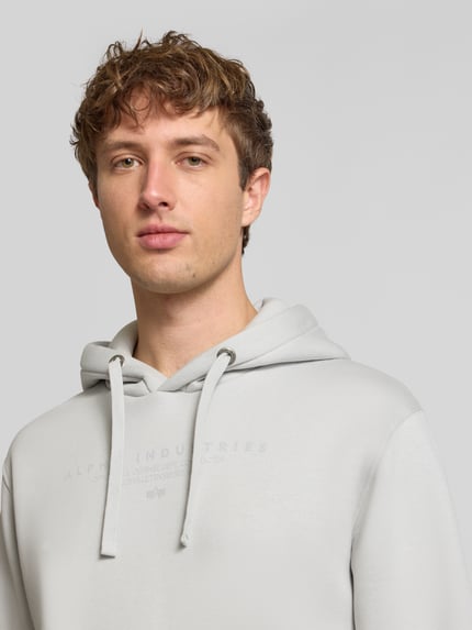 Männermodel trägt Relaxed Fit Hoodie aus Baumwoll-Mix mit Label-Detail von Alpha Industries, hellgrau, Kapuze, Nahaufnahme Oberkörper.