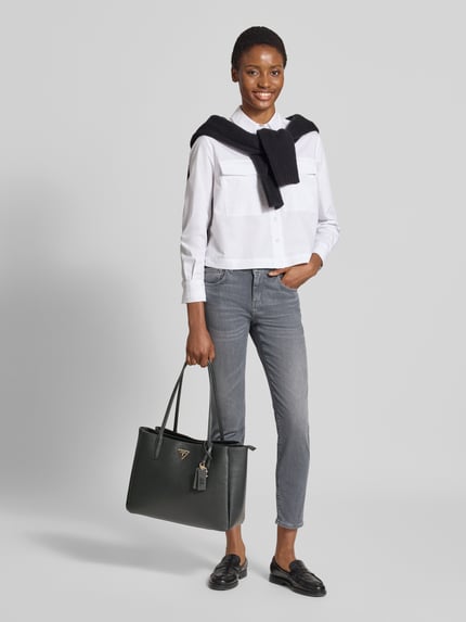 Vrouwenmodel draagt Tote Bag in Leder-Optik model 'Noelle' van GUESS in zwart met logodetail, smalle schouderband, magneetsluiting, getailleerd hoofdvak, Breedte 39 cm, Lengte 28 cm, Diepte 14 cm, bandlengte 64 cm, leerlook. Volledige lichaamsaanzicht van voren.