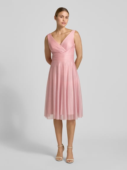 Frauenmodel trägt Knielanges Cocktailkleid mit V-Ausschnitt von TROYDEN COLLECTION in zartem Rosa, Chiffon, taillierter Schnitt, Ganzkörperansicht von vorne.