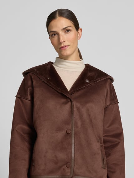 Frauenmodel trägt Jacke mit Kapuze und seitlichen Eingrifftaschen von Apricot, braun, weiche Textur, Nahaufnahme Oberkörper, gerader Schnitt, Druckknopfverschluss, KI-modifiziertes Bild.